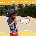 Child holding a toy camera with a speech bubble saying 'Smile! You're in my photo!' against a natural background.