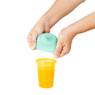 Child's hands using a green lid to open a yellow cup filled with orange juice on a white background