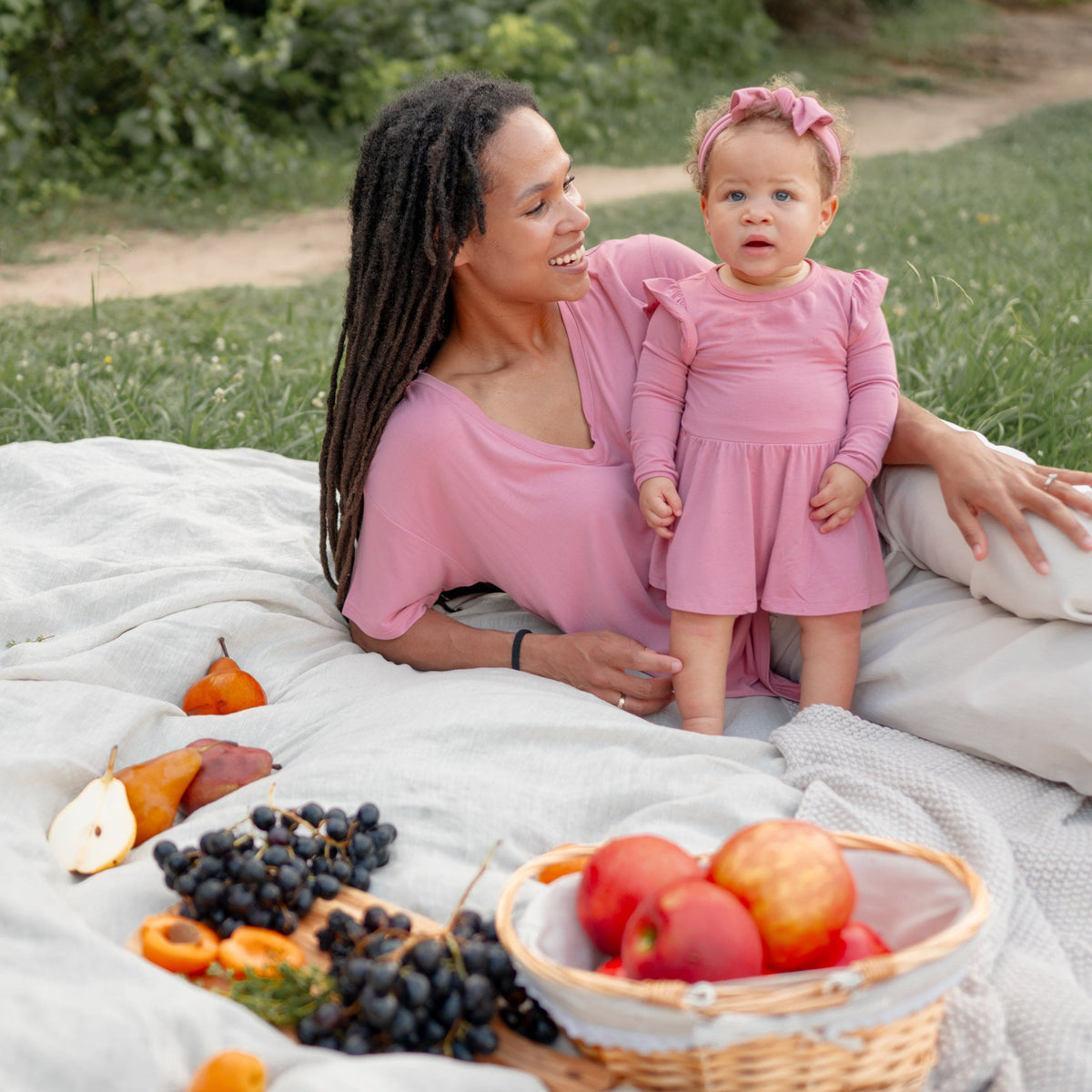 Woman and child in pink outfits sitting on a blanket with fruits around them outdoors.
