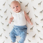 Baby lying on a patterned surface wearing a white sleeveless shirt and blue pants.