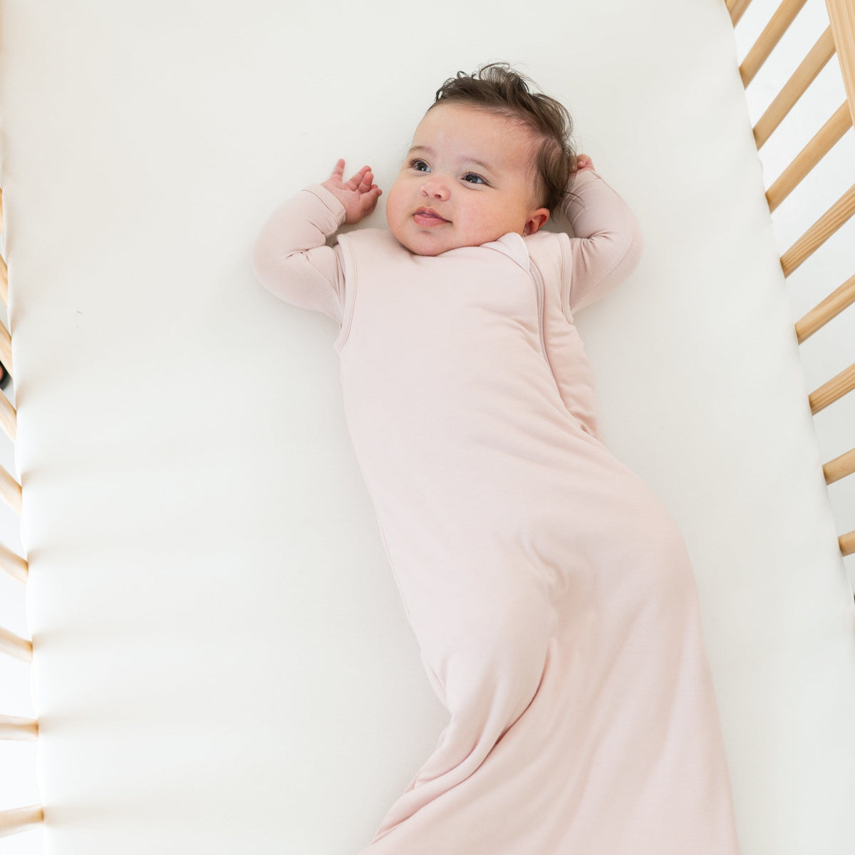 Baby wrapped in a pink blanket lying on a white surface with wooden crib bars.