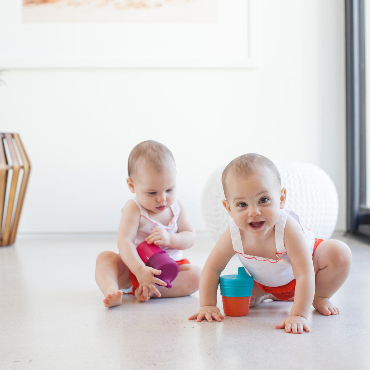 Two babies playing on a light floor in a bright room, one sitting and holding a purple cup, and one crawling while holding a teal and orange cup. Both wear matching white tank tops with orange shorts.