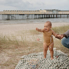 Baby in a swimsuit standing on a blanket with a beach and pier in the background