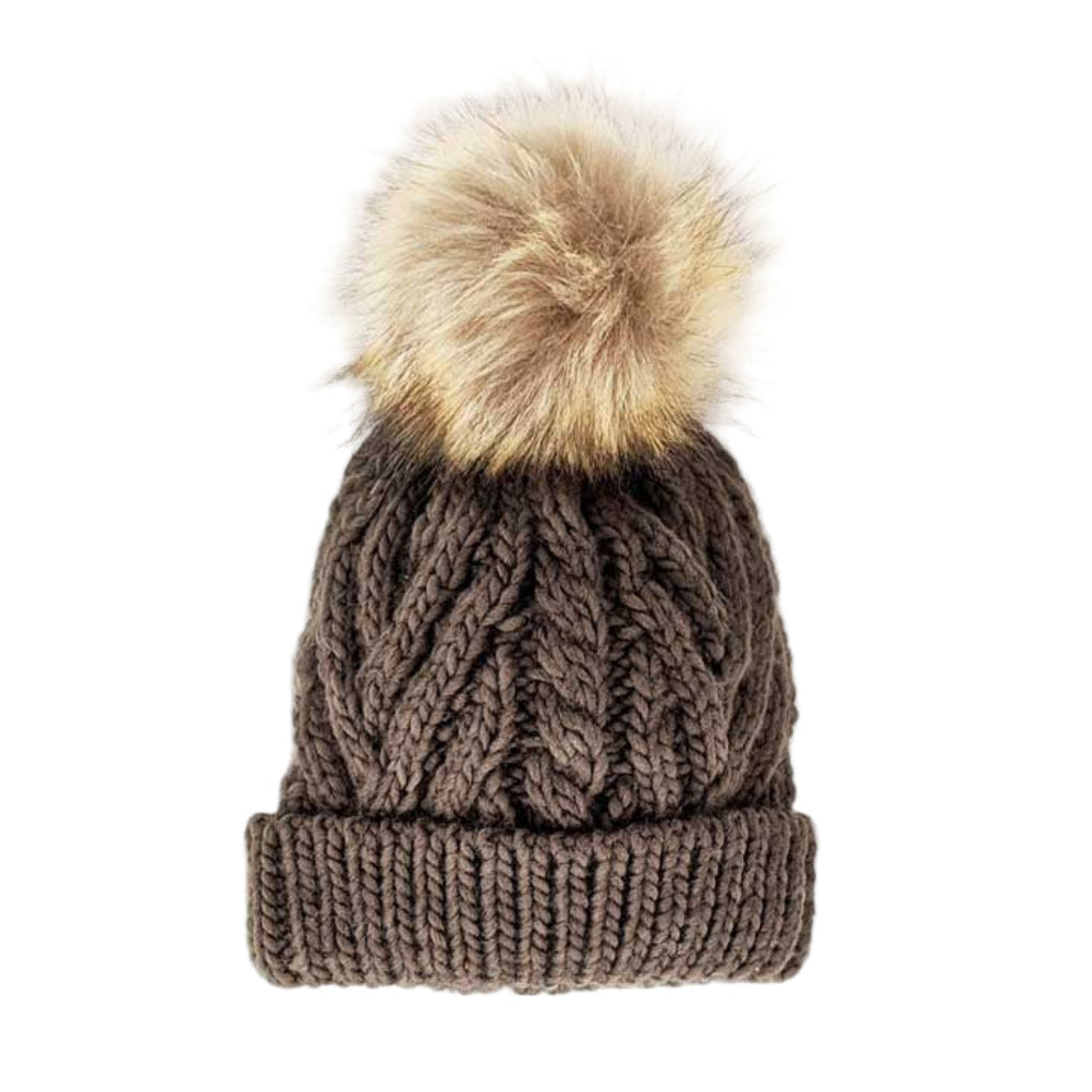 A brown cable-knit beanie hat with a ribbed cuff and a large fluffy beige faux-fur pom-pom on top, shown against a plain white background.