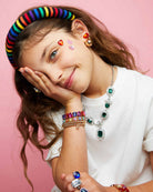 Young girl with colorful jewelry including a rainbow headband, necklaces, bracelets, and rings on a pink background.
