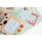 Children's drawing board with colorful shapes and a small book on a light green surface.