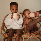 Two children sitting on a wicker chair, drinking from white cups.
