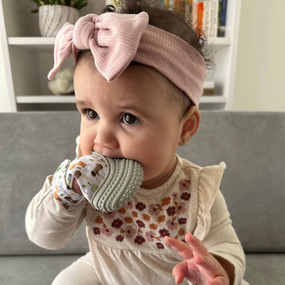 Baby wearing a pink headband with bow and holding a teething ring indoors.
