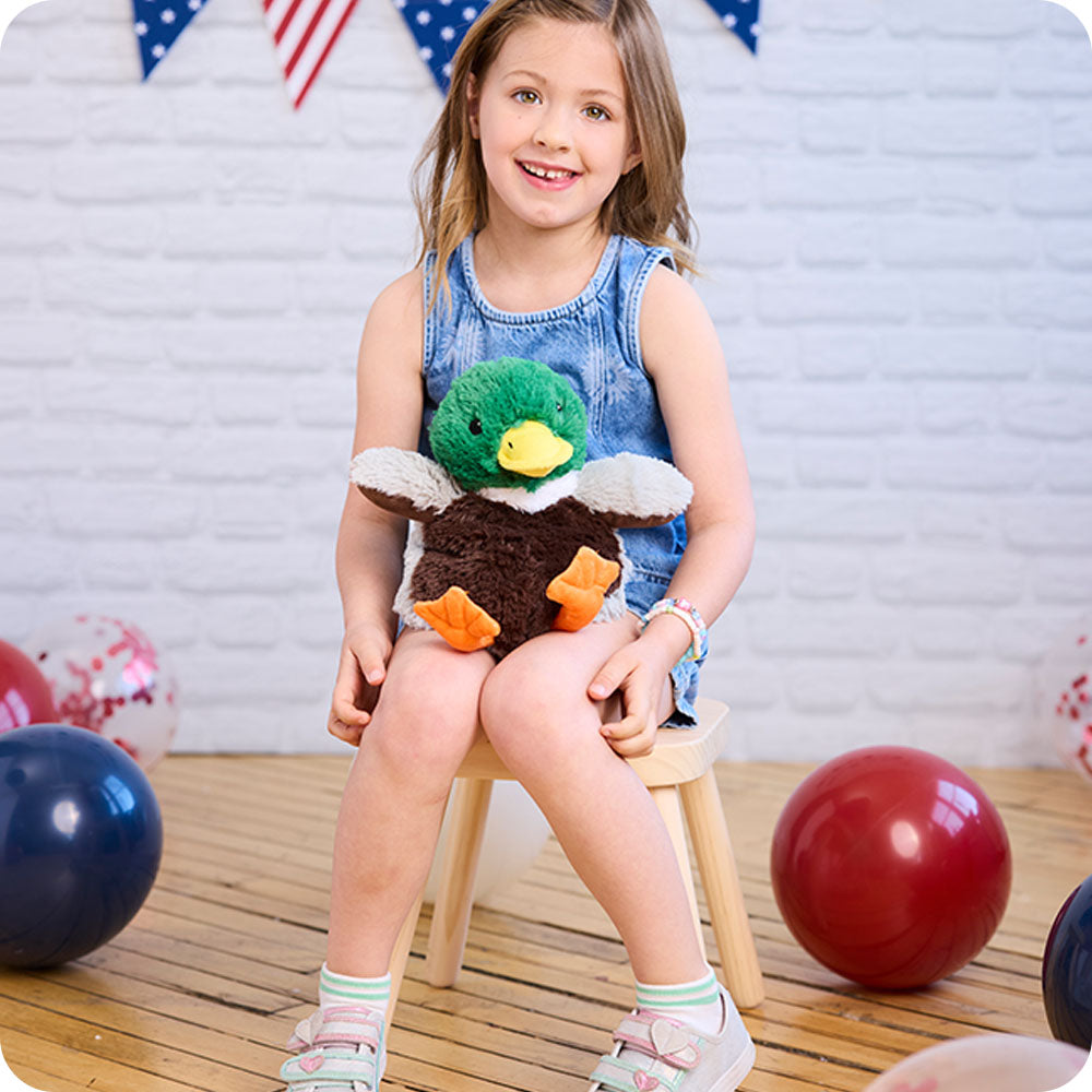 Young girl sitting on a chair holding a green plush duck toy with balloons in the background.