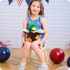 Young girl sitting on a chair holding a green plush duck toy with balloons in the background.