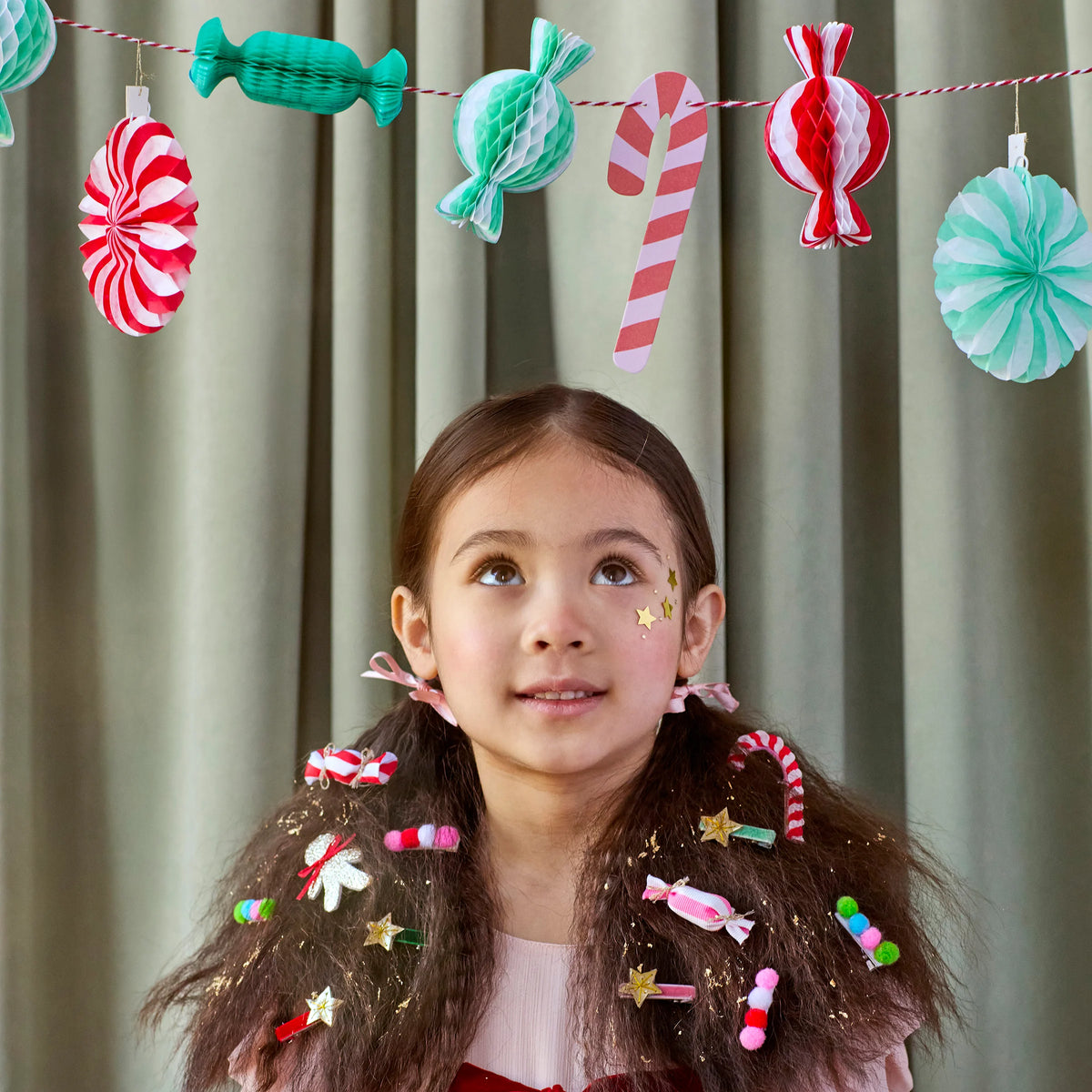 A young girl wearing the festive Meri Meri Gingerbread House Hair Clips set of 9 whimsical clips featuring candy and gingerbread designs, with a matching Christmas Candy Mini Honeycomb Garland hanging in the background.
