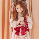 A young girl with long red hair wearing a pale pink dress and decorative hair clips, holding a small shiny pink bag with a large red bow and red strap, posed in front of peach-colored curtains.