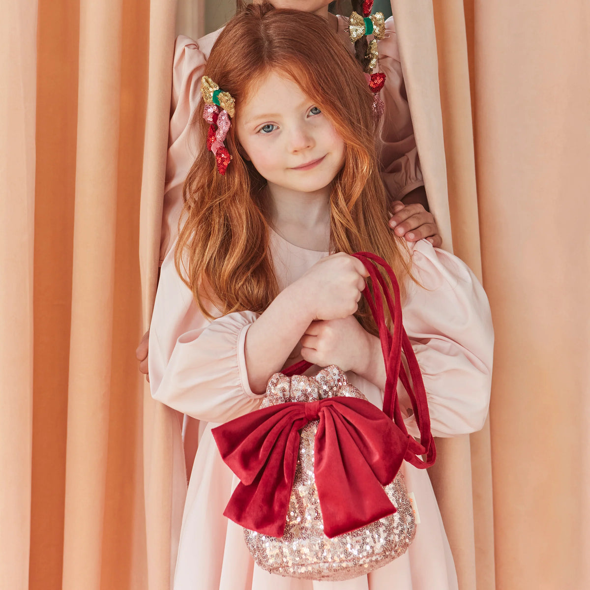 A young girl with long red hair wearing a pale pink dress and decorative hair clips, holding a small shiny pink bag with a large red bow and red strap, posed in front of peach-colored curtains.