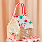 A straw house-shaped handbag with a red and white striped handle, decorated with candy canes, colorful pom-poms, and white lace-like trim, set against a soft peach curtain background.
