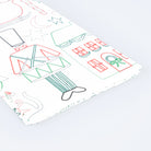 A folded sheet of white paper with a whimsical line-art holiday illustration pattern, featuring a nutcracker, house, stars, and ornaments in red, green, and gray, photographed on a clean white background.