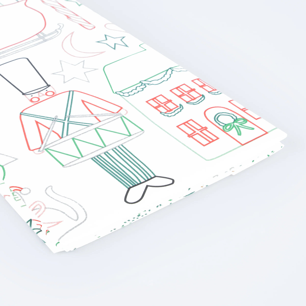 A folded sheet of white paper with a whimsical line-art holiday illustration pattern, featuring a nutcracker, house, stars, and ornaments in red, green, and gray, photographed on a clean white background.