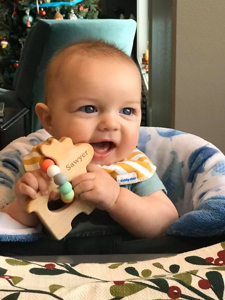 Baby holding a wooden toy with 'Sawyer' on it, sitting in a high chair.