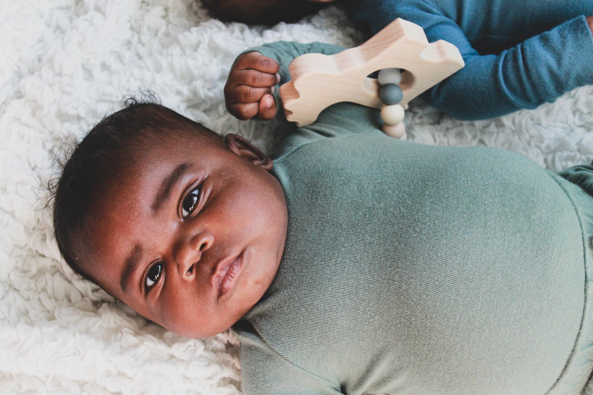 Baby lying on a white surface with a toy, wearing a green outfit.