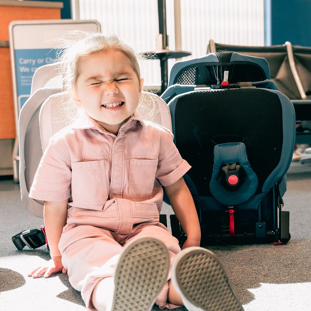 Child in a pink outfit sitting next to a car seat in an indoor setting
