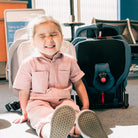 Child in a pink outfit sitting next to a car seat in an indoor setting