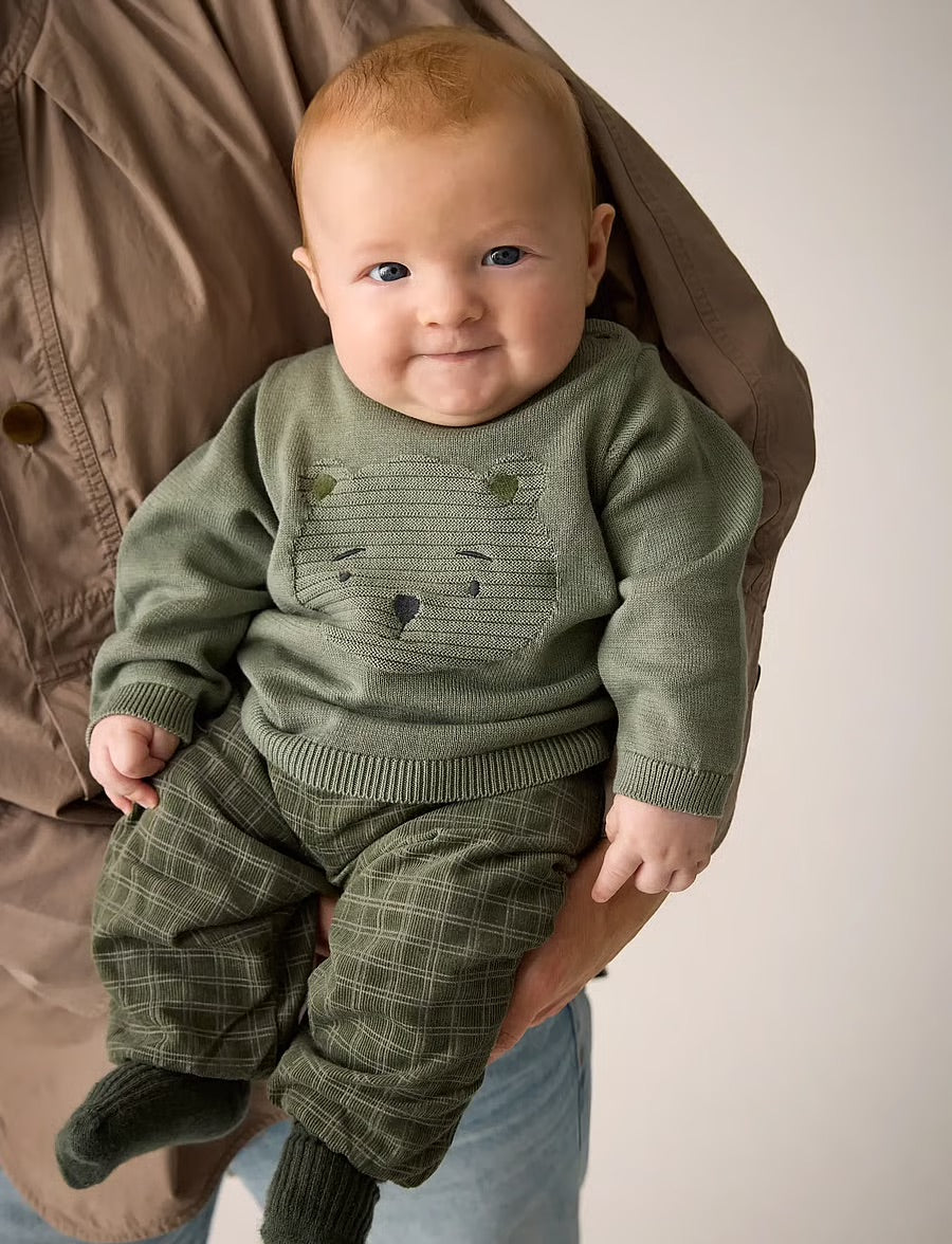 A baby wearing an olive green knit sweater and matching plaid pants, held by an adult wearing a brown jacket against a plain light background.