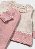 A baby knit sweater in cream and pink, featuring textured knit patterns, a scalloped collar, and a matching pink knit accessory partially draped over the front.