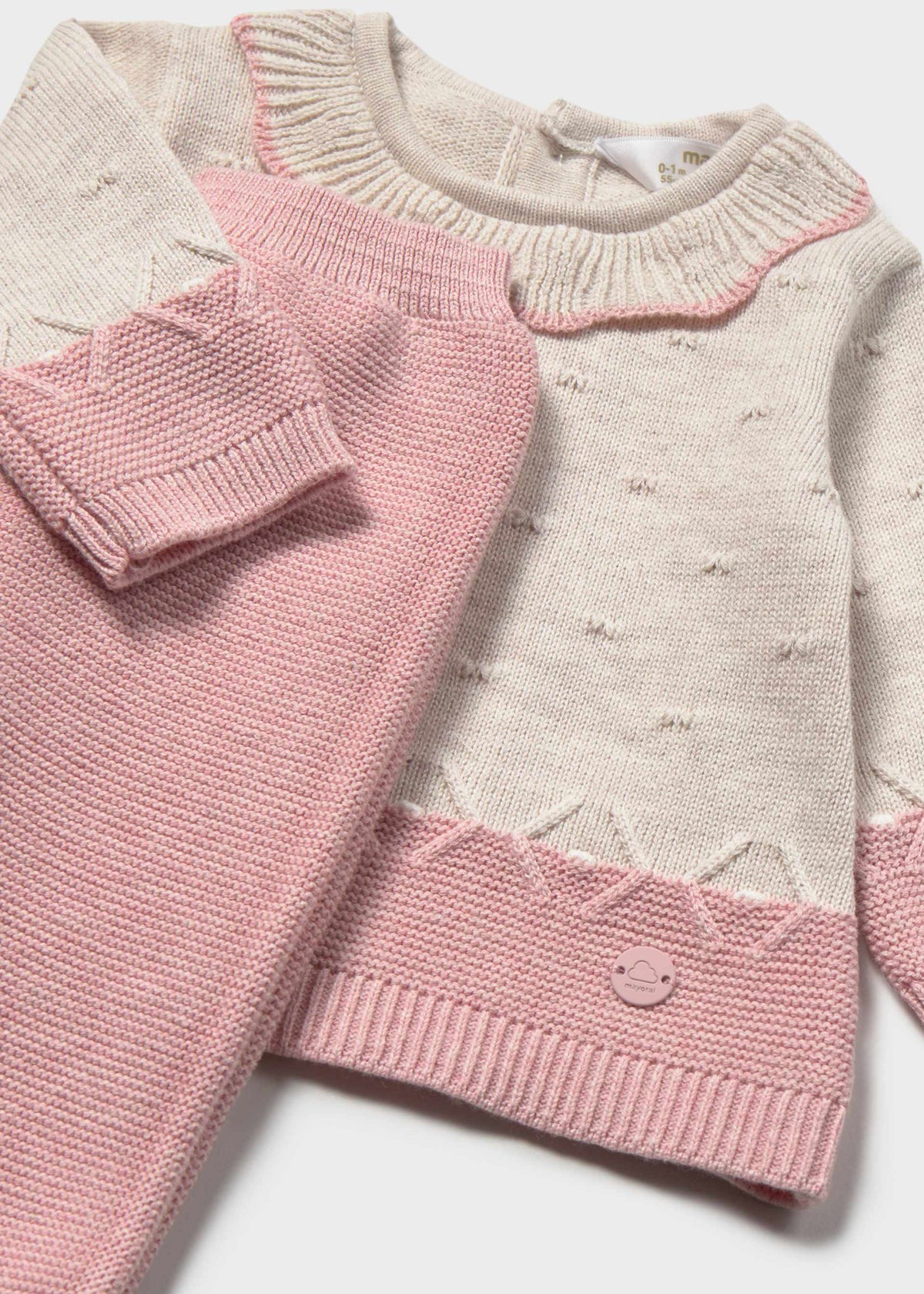 A baby knit sweater in cream and pink, featuring textured knit patterns, a scalloped collar, and a matching pink knit accessory partially draped over the front.