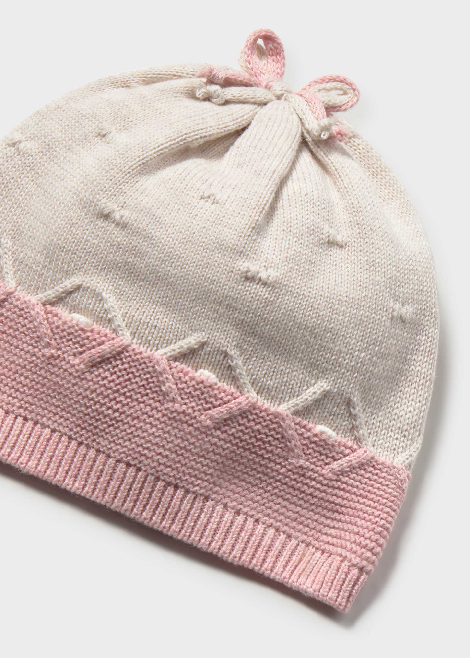 A close-up of a knitted baby hat in off-white with a pink ribbed brim and a matching pink drawstring bow at the top, shown on a light gray background.