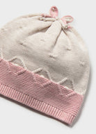 A close-up of a knitted baby hat in off-white with a pink ribbed brim and a matching pink drawstring bow at the top, shown on a light gray background.