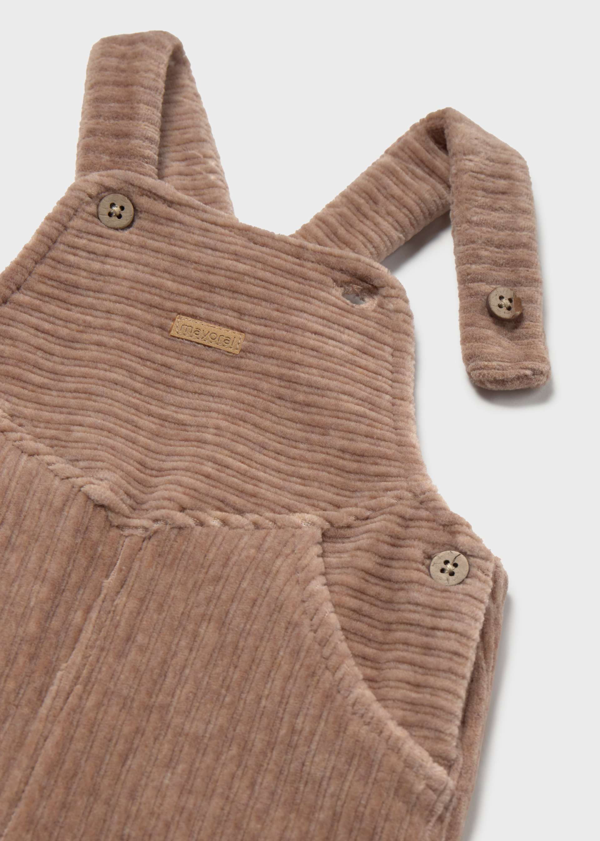 A close-up of a child's corduroy overall in a warm taupe-brown color, featuring adjustable shoulder straps with decorative buttons and a small brand label.