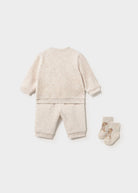 A baby loungewear set consisting of a long-sleeve sweatshirt and matching pants in a light beige heathered fabric, shown from the back next to a pair of matching socks on a light gray background.