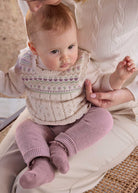 Baby wearing a knitted outfit with a patterned sweater and pink pants, sitting on a cushion.
