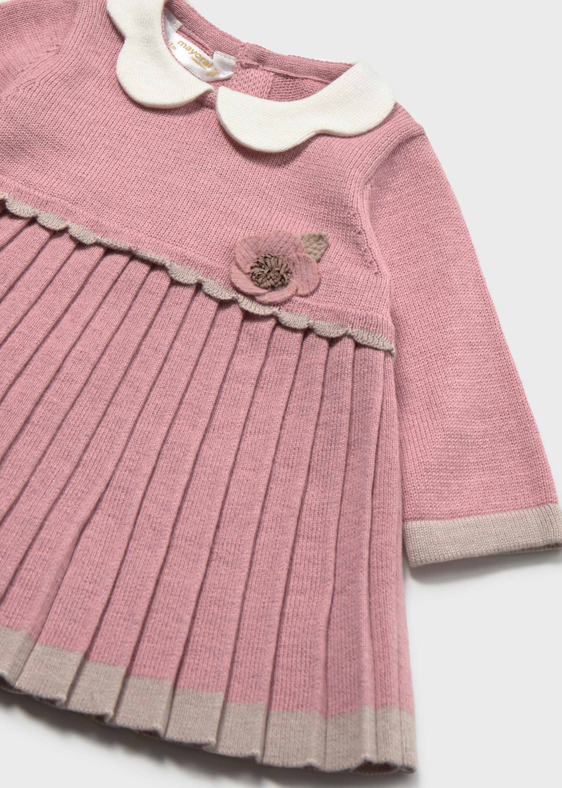 A close-up of a pink ribbed knit dress for a young child, featuring a white Peter Pan collar, a pleated skirt, and a decorative fabric flower on the chest.