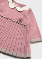 A close-up of a pink ribbed knit dress for a young child, featuring a white Peter Pan collar, a pleated skirt, and a decorative fabric flower on the chest.
