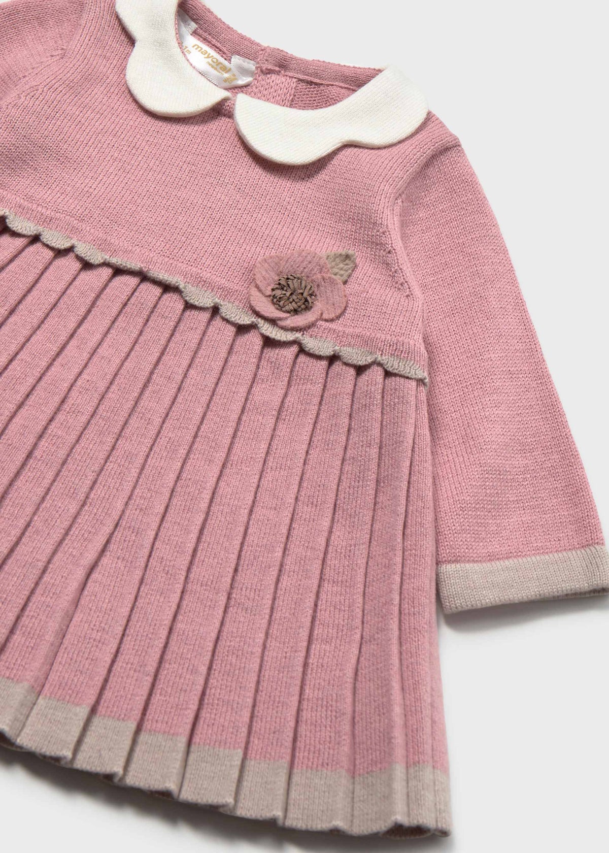 A close-up of a pink ribbed knit dress for a young child, featuring a white Peter Pan collar, a pleated skirt, and a decorative fabric flower on the chest.