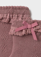 Close-up of a dusty pink knit garment with ruffled trim and a matching ribbon bow, shown against a light neutral background.