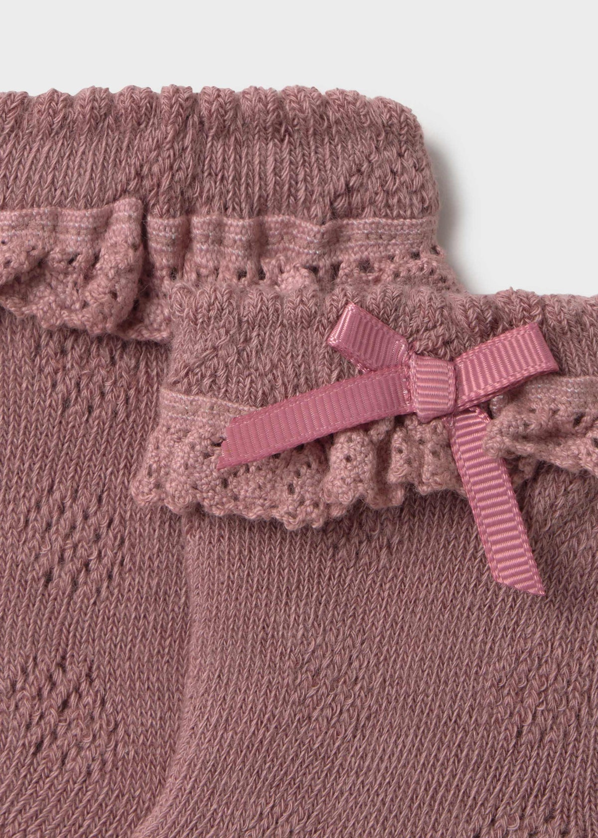 Close-up of a dusty pink knit garment with ruffled trim and a matching ribbon bow, shown against a light neutral background.