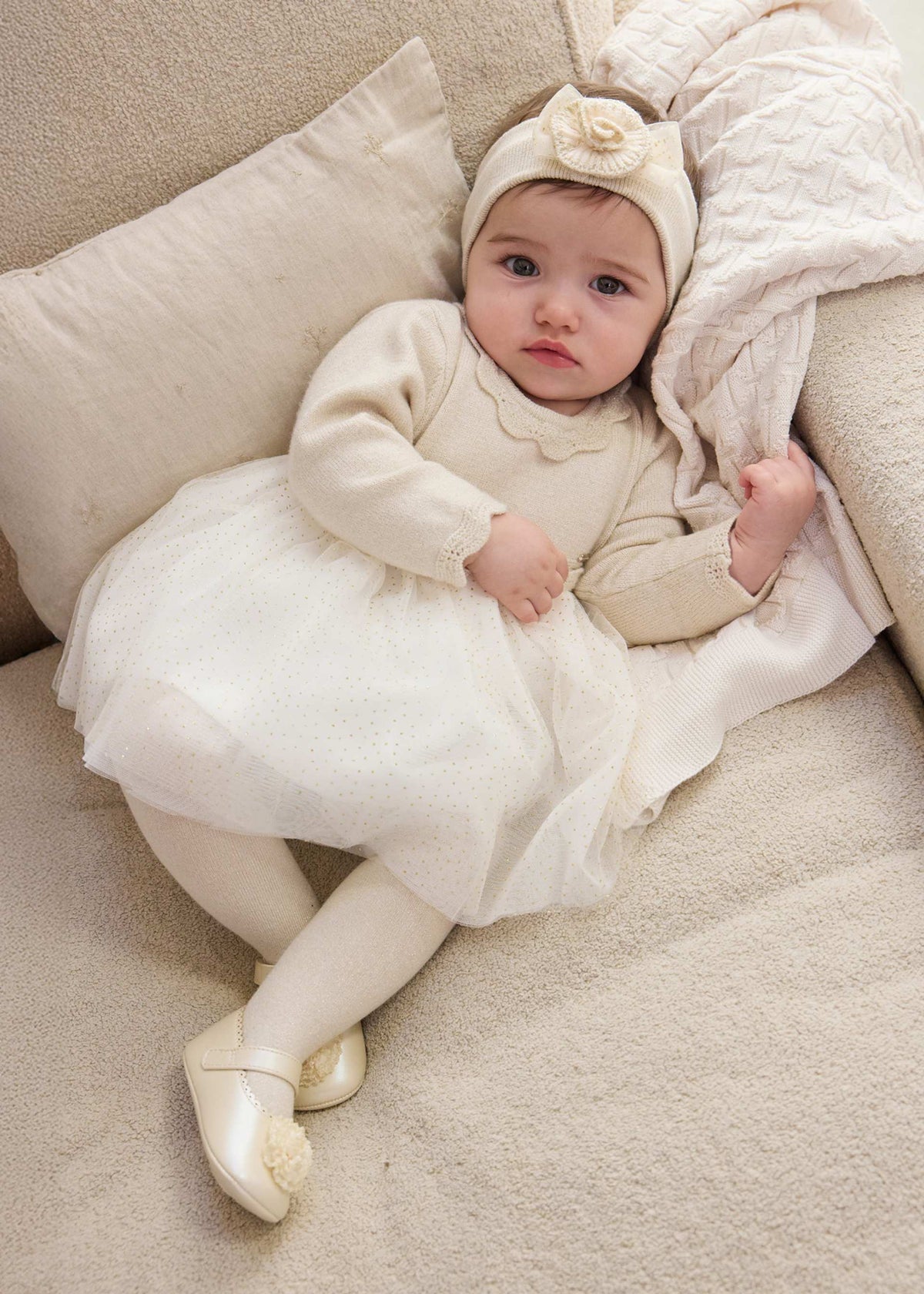 Baby in a white outfit and headband lying on a beige couch.