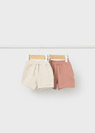 Two pairs of shorts, one beige and one pink, hanging on a white background.