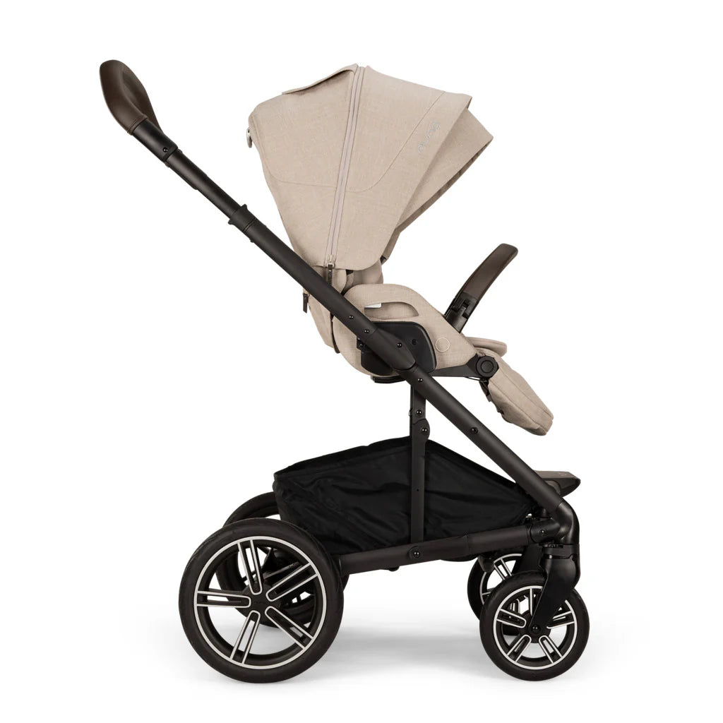 A profile view of a MIXX Next + Pipa Aire RX Travel System by Nuna stroller in Biscotti featuring a beige seat and canopy, a black frame, and a brown leatherette handlebar and armbar, set against a clean white background.