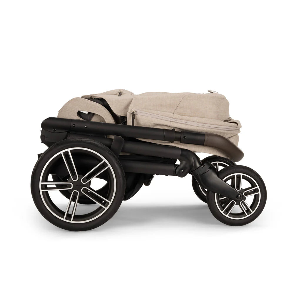 A profile view of a folded MIXX Next + Pipa Aire RX Travel System by Nuna stroller in Biscotti featuring a compact fold with an exposed brown leatherette handlebar, set against a clean white background.