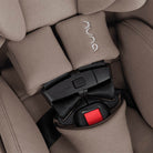 Close-up of a Nuna car seat with beige fabric padding and a black buckle featuring a red release button. The Nuna logo is embroidered on the headrest and embossed on the buckle.