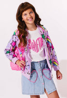 Young girl wearing a colorful jacket, white shirt with pink text, and denim skirt with heart designs on a light background.