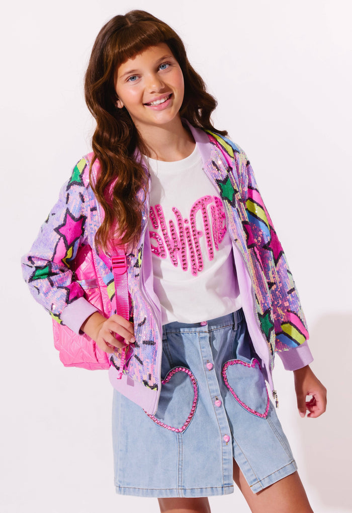 Young girl wearing a colorful jacket, white shirt with pink text, and denim skirt with heart designs on a light background.