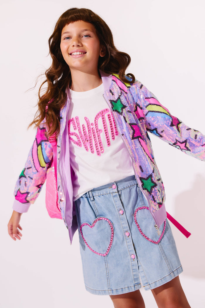 Young girl wearing a colorful jacket, white shirt with pink text, and denim skirt with heart designs on a light background.