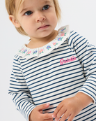 A close-up of a young child wearing a navy and white horizontally striped long-sleeve top with a white embroidered floral collar and a pink 'Gabbie' script on the chest, against a light gray background.