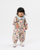 A toddler girl wearing a long-sleeve floral jumpsuit with a light blue ruffled collar, standing against a plain white studio background.