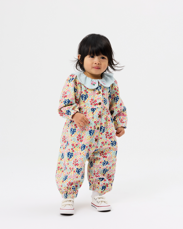 A toddler girl wearing a long-sleeve floral jumpsuit with a light blue ruffled collar, standing against a plain white studio background.