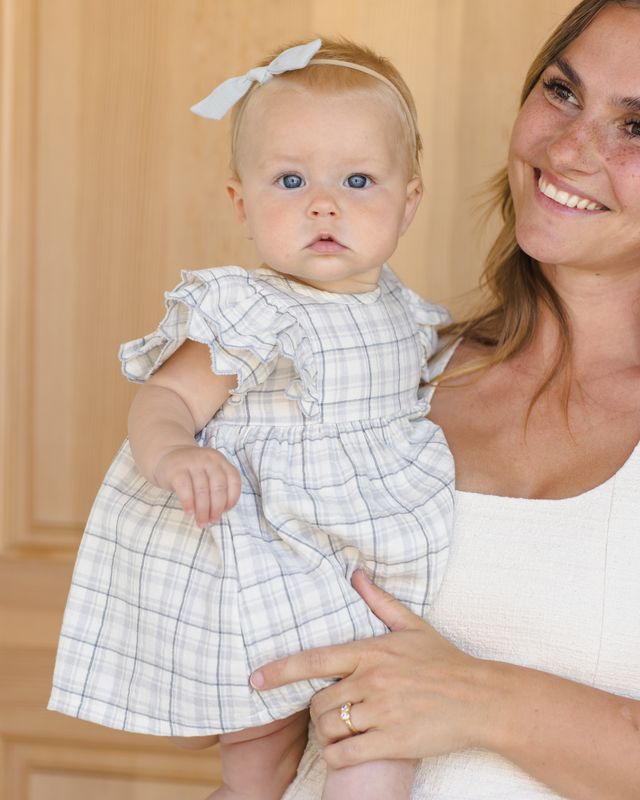 Woman holding a baby wearing a checkered dress indoors.