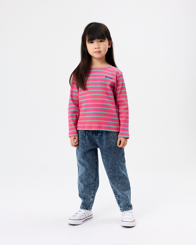 A young girl stands against a plain white studio background wearing a pink and light blue horizontally striped long-sleeve knit top, blue jeans, and white sneakers.